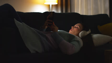 Woman-Wearing-Wireless-Headphones-Lying-On-Sofa-At-Home-At-Night-Streaming-Or-Looking-At-Online-Content-On-Mobile-Phone-4